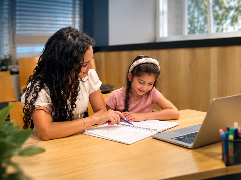 Een begeleider van Sterren van Straks helpt een jong meisje met haar schoolwerk aan een tafel met een schrift en een laptop.